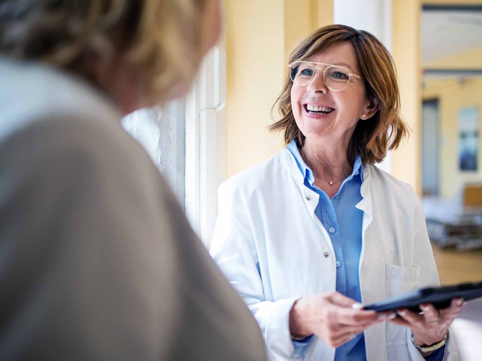 A practitioner holds a tablet computer while smiling and talking to a patient in medical facility