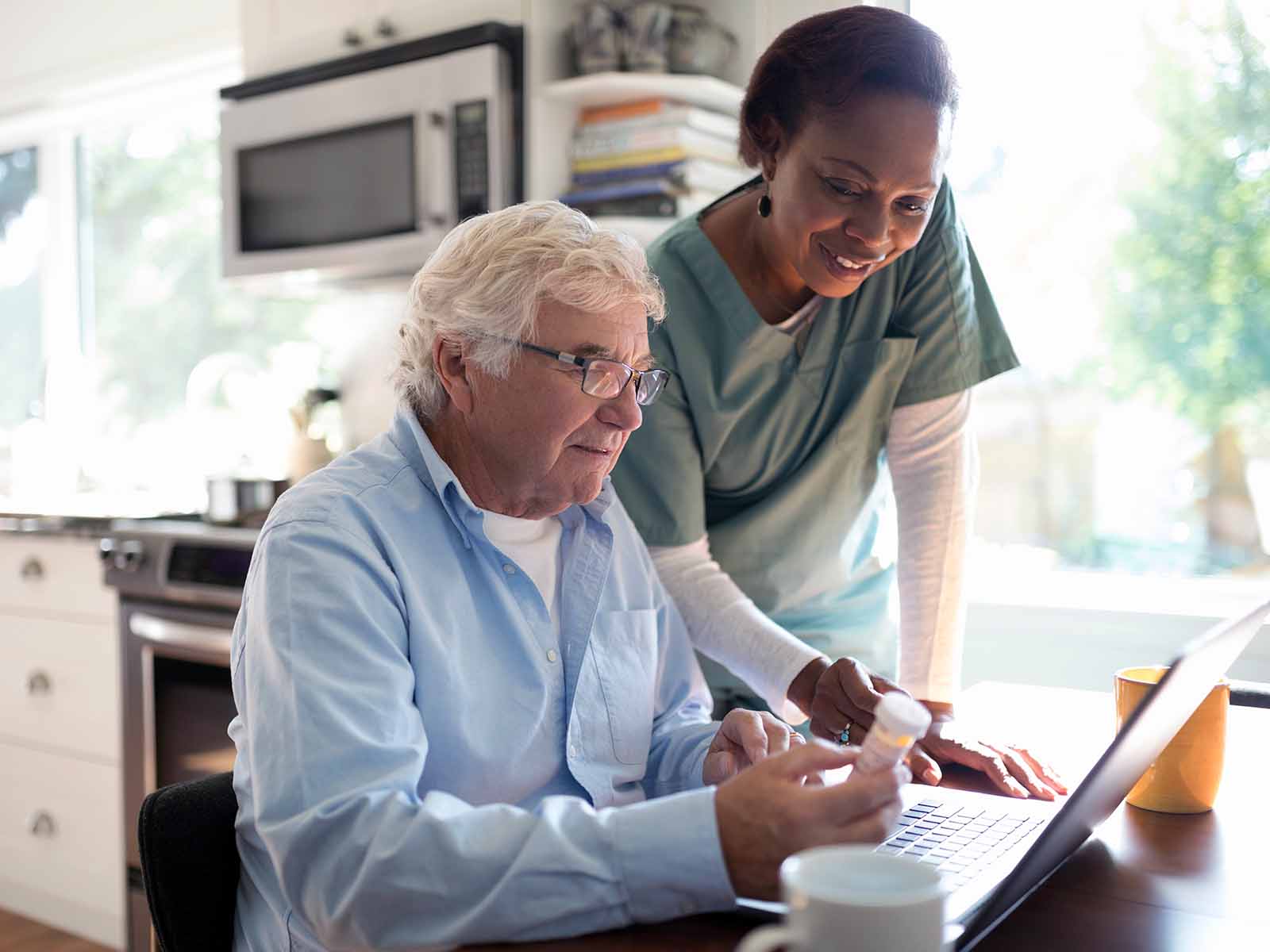 An older male patient reorders prescriptions online, assisted by an in-home health professional